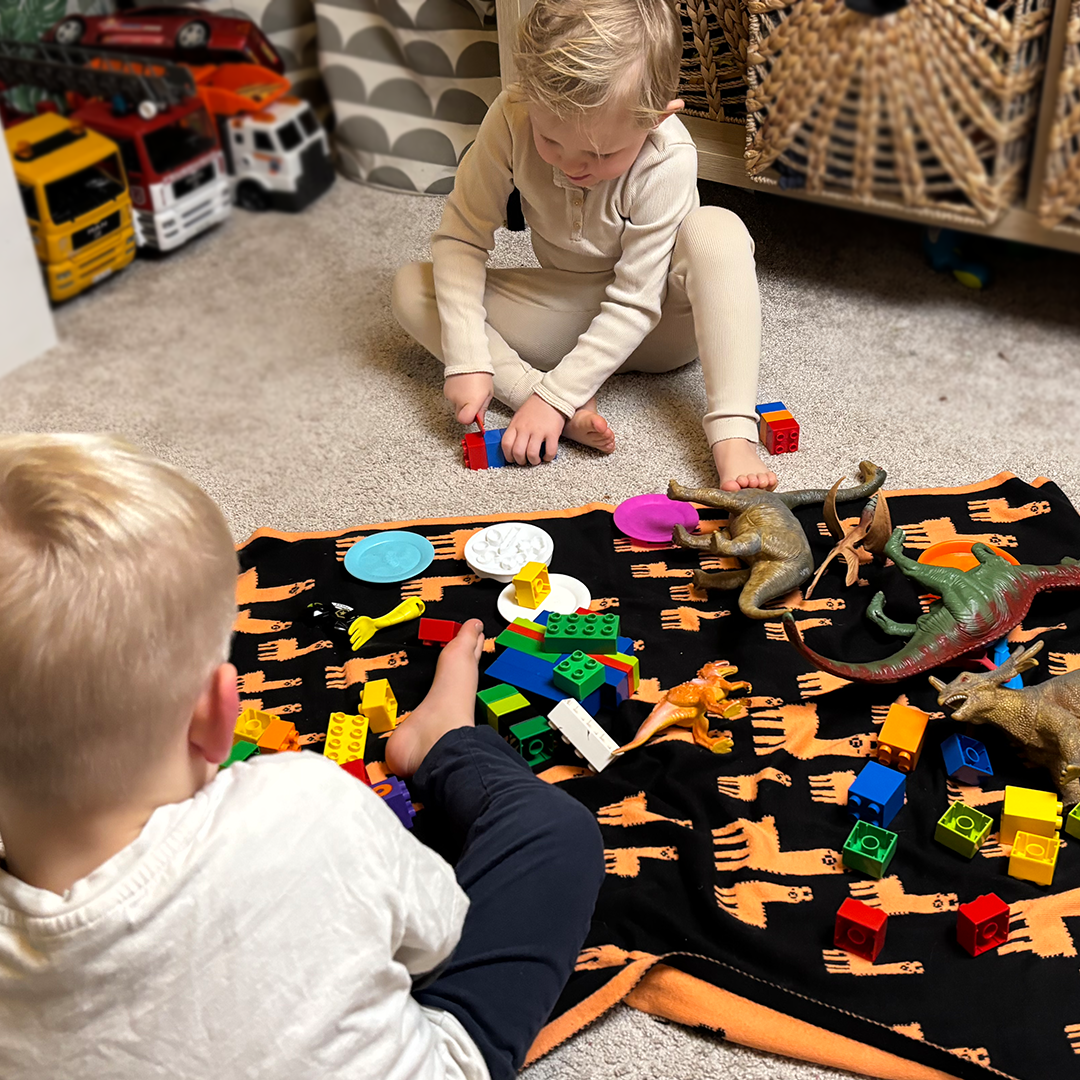 barn som sitter på golvet och har dukat upp en picknick med lego och dinosaurier på en färgglad filt